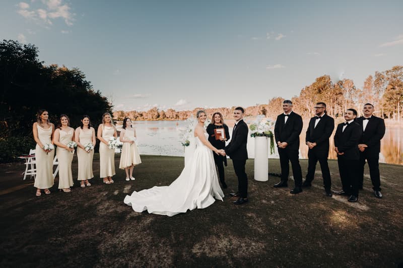Bride Libby and groom Kyle stand holding hands at the ceremony stage at The Tides — The Water's Edge, with the officiant between them. Bridesmaids in matching cream dresses hold bouquets on the left, and groomsmen in black tuxedos stand on the right, with a lake and trees in the background.