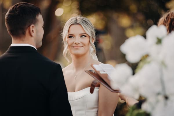 Bride Libby and groom Kyle face each other during their wedding ceremony at The Tides — The Water's Edge, with an officiant holding a book partially visible in the foreground.