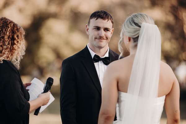 Kyle in a black tuxedo and bow tie faces Libby in a white strapless wedding gown with a veil during their ceremony at The Tides — The Water's Edge, while an officiant holds a microphone and papers nearby.
