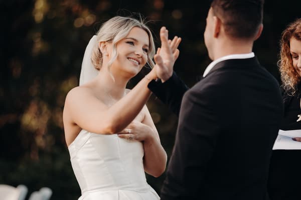 Bride Libby and groom Kyle exchange a hand gesture during their wedding ceremony at The Tides — The Water's Edge, with the officiant partially visible holding papers.
