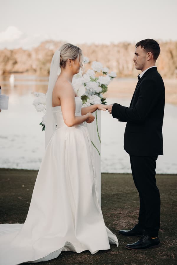 Bride Libby and groom Kyle exchange rings during their wedding ceremony at The Tides — The Water's Edge, standing outdoors near a body of water with floral arrangements behind them.