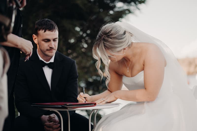 The bride Libby signs a document at a small table while the groom Kyle sits beside her at The Tides — The Water's Edge during the ceremony.