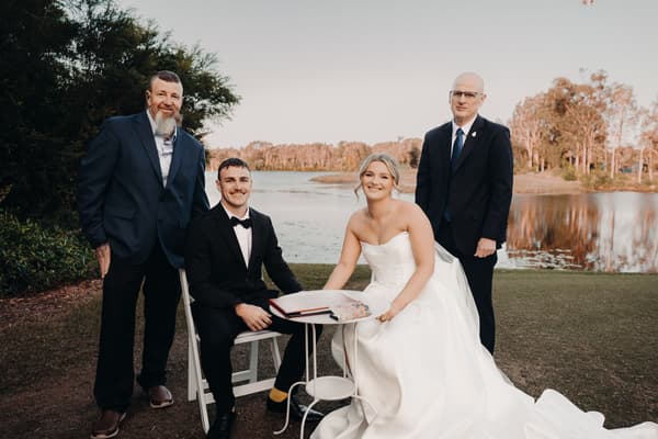 Bride Libby in a white wedding gown and groom Kyle in a black tuxedo sit at a small table signing a book outdoors at The Tides — The Water's Edge, flanked by two older men in dark suits standing on either side, with a lake and trees in the background.