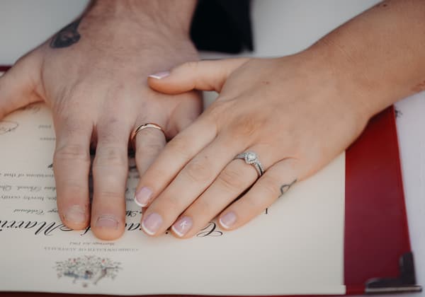 Close-up of the bride and groom's hands resting on a marriage certificate at The Tides — The Water's Edge, showing their wedding rings.
