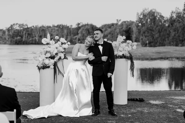 Bride Libby and groom Kyle stand together at the ceremony stage by the water's edge at The Tides, with floral arrangements on white pedestals behind them.