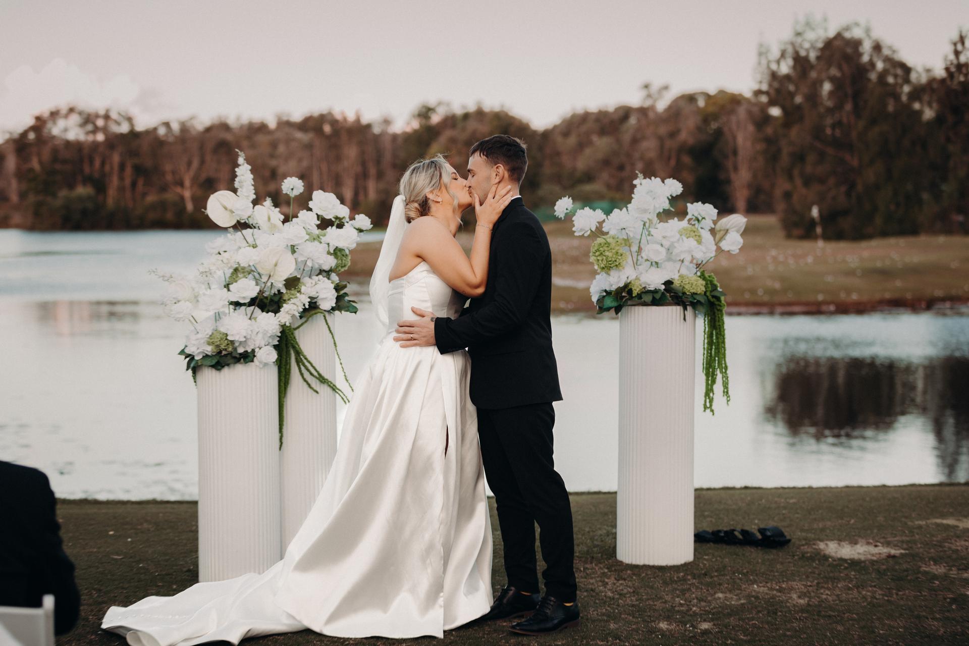 Bride Libby and groom Kyle share a kiss at the ceremony stage by the water's edge at The Tides, flanked by two tall white floral arrangements.