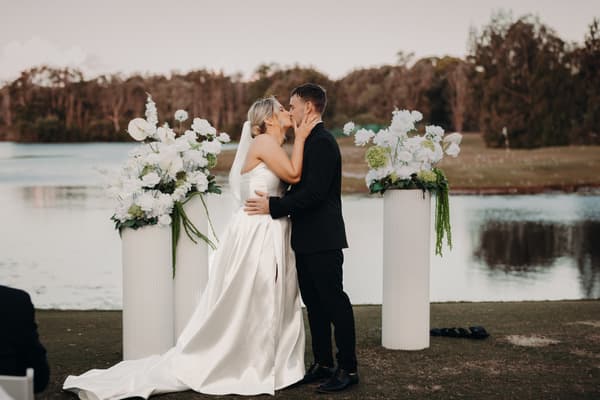 Bride Libby and groom Kyle share a kiss at the ceremony stage by the water's edge at The Tides, flanked by two tall white floral arrangements.