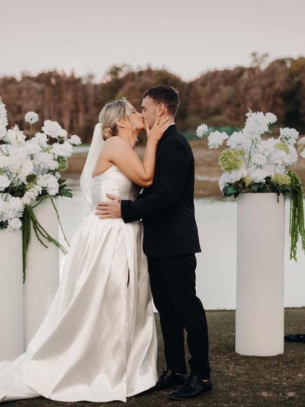 Bride Libby and groom Kyle share a kiss at the ceremony stage by the water's edge at The Tides, flanked by two tall white floral arrangements.