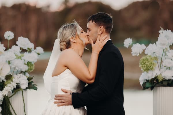 Bride Libby and groom Kyle share a kiss at the ceremony stage at The Tides — The Water's Edge, surrounded by white floral arrangements.