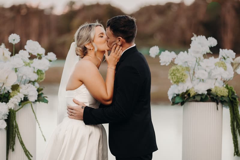Bride Libby and groom Kyle share a kiss at the ceremony stage at The Tides — The Water's Edge, flanked by white floral arrangements.