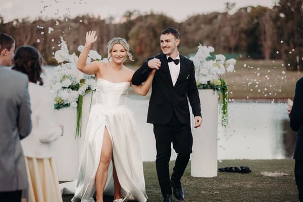 Bride Libby and groom Kyle walk hand in hand at The Tides — The Water's Edge ceremony stage, with Libby waving to guests as white confetti falls around them.
