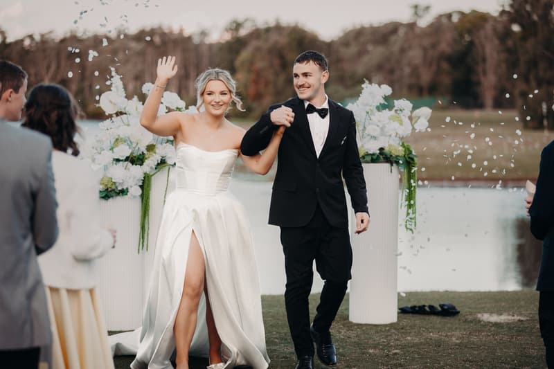 Bride Libby and groom Kyle walk hand in hand at The Tides — The Water's Edge ceremony stage, with Libby waving to guests as white confetti falls around them.