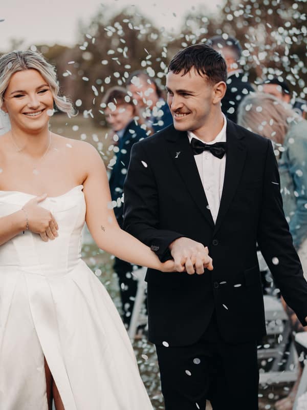 Bride Libby and groom Kyle walk hand in hand outdoors at The Tides — The Water's Edge, surrounded by guests and falling confetti.