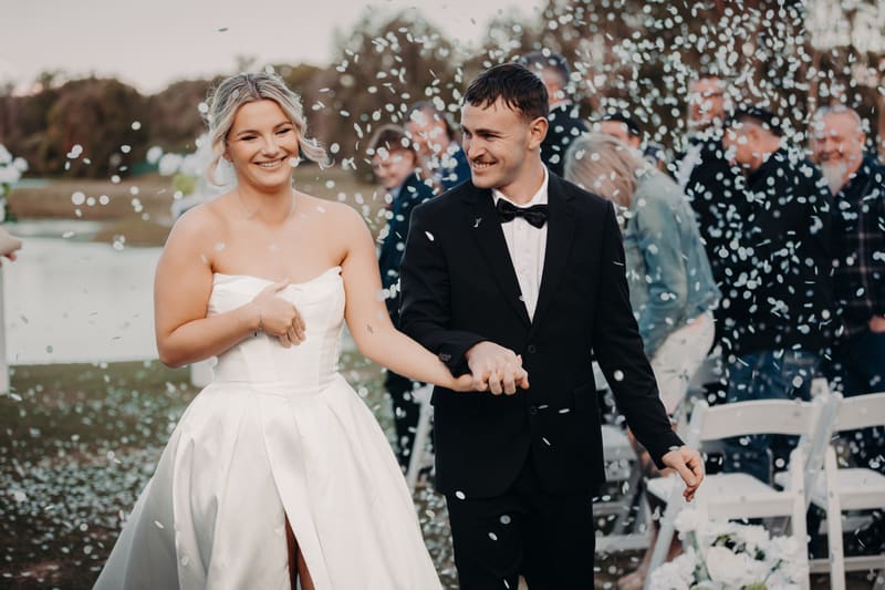 Bride Libby and groom Kyle walk hand in hand outdoors, surrounded by guests and falling confetti. Best time of year to get married by She Said Yes.