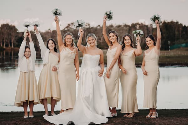 Libby the bride stands with six bridesmaids and flower girl outdoors by a lake, all holding bouquets raised above their heads. Cinematic photography wedding by She Said Yes.