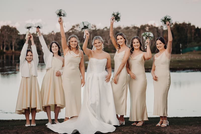 Libby the bride stands with six bridesmaids and flower girl outdoors by a lake at The Tides, all holding bouquets raised above their heads.