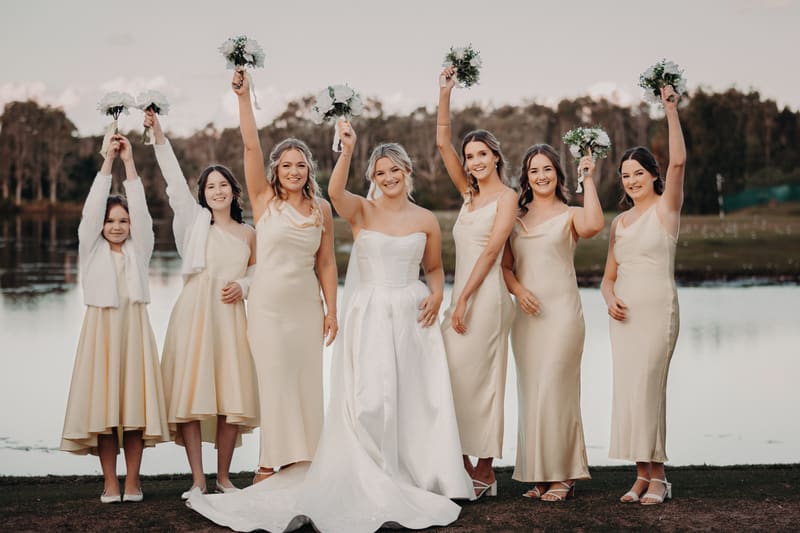 Libby the bride stands with six bridesmaids and flower girl outdoors by a lake, all holding bouquets raised above their heads. Cinematic photography wedding by She Said Yes.