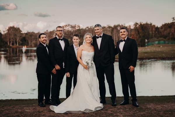 Libby the bride in a white strapless gown holding a bouquet stands with Kyle the groom and four groomsmen dressed in black tuxedos near a lake. Wedding industry awards finalist by She Said Yes.