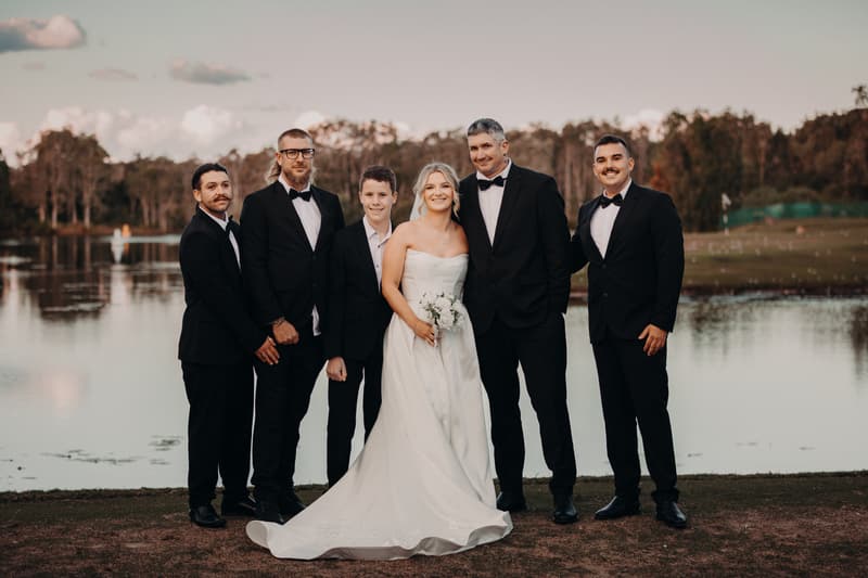 Libby the bride in a white strapless gown holding a bouquet stands with Kyle the groom and four groomsmen dressed in black tuxedos near a lake at The Tides.