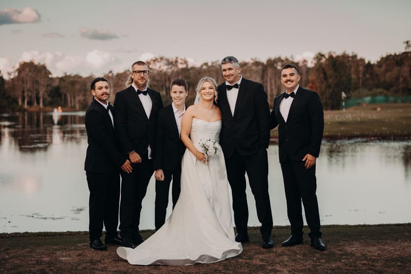 Libby the bride in a white strapless gown holding a bouquet stands with Kyle the groom and four groomsmen dressed in black tuxedos near a lake. Wedding industry awards finalist by She Said Yes.