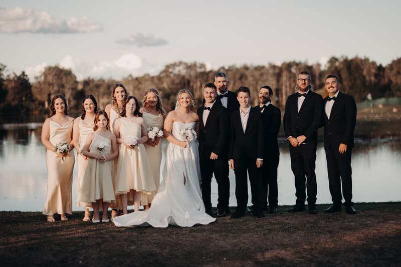 Libby the bride in a white gown and Kyle the groom in a black tuxedo stand with their bridal party dressed in cream dresses and black tuxedos near a lake at The Tides.