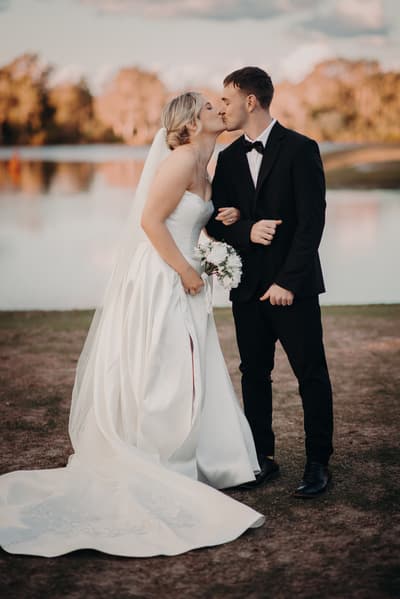 Bride Libby and groom Kyle share a kiss during their couple portraits session at The Tides, standing outdoors near a body of water with trees in the background. Libby wears a strapless white wedding gown with a long train and holds a bouquet, while Kyle is dressed in a black tuxedo with a bow tie.