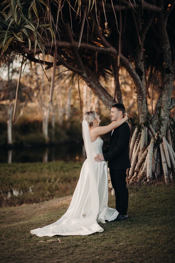 Bride Libby and groom Kyle stand embracing under a large tree at The Tides during their couple portraits session. Libby wears a white wedding gown with a long train and veil, while Kyle is dressed in a black suit.