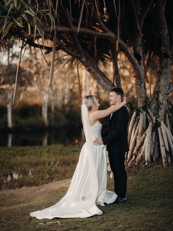 Bride Libby and groom Kyle stand embracing under a large tree at The Tides during their couple portraits session. Libby wears a white wedding gown with a long train and veil, while Kyle is dressed in a black suit.