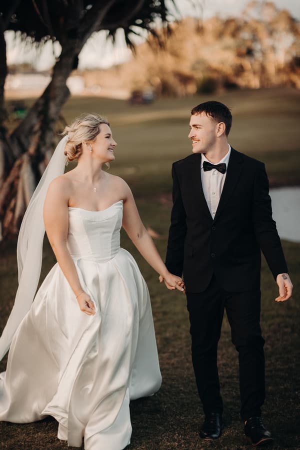 Libby in a strapless white wedding gown and veil holding hands with Kyle in a black tuxedo with a bow tie, walking outdoors at The Tides.
