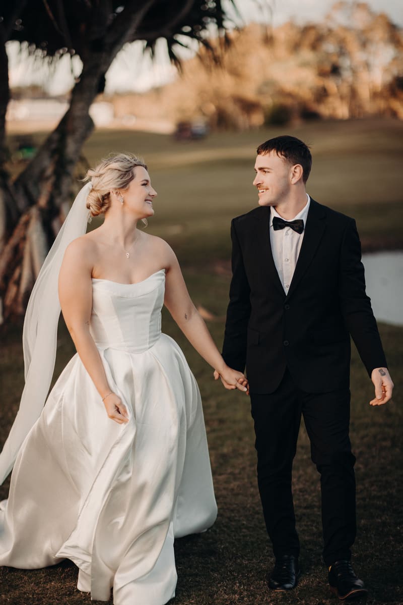 Libby in a strapless white wedding gown and veil holding hands with Kyle in a black tuxedo with a bow tie, walking outdoors. Wedding photography coverage options by She Said Yes.