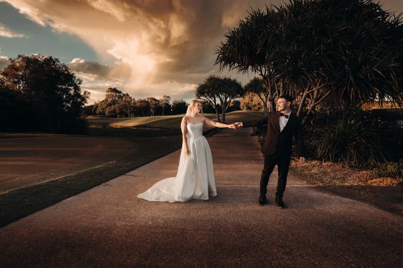 Bride Libby and groom Kyle hold hands while standing on a paved path at The Tides, surrounded by trees and greenery under a dramatic cloudy sky.