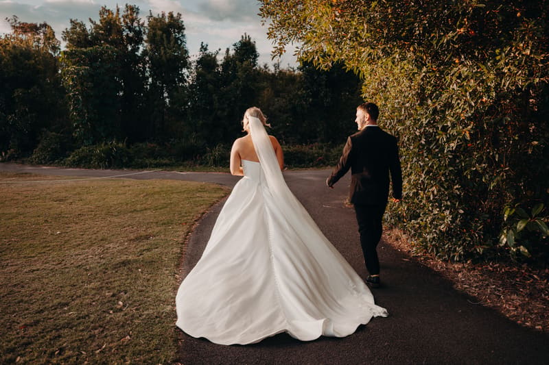 The bride Libby and the groom Kyle walk side by side on a paved path surrounded by greenery, with Libby wearing a strapless wedding gown and veil and Kyle in a dark suit.