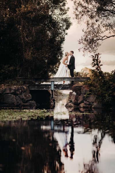 Bride Libby and groom Kyle stand on a small wooden bridge over a reflective body of water, surrounded by trees and natural foliage, sharing a kiss. Outdoor photo booth hire by She Said Yes.