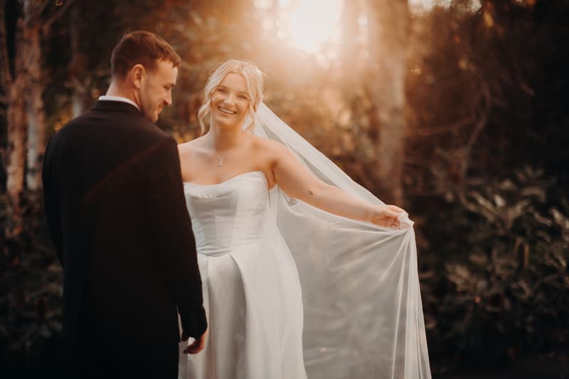 Bride Libby and groom Kyle pose together outdoors at The Tides during their couple portraits session, with Libby holding out her veil and smiling.