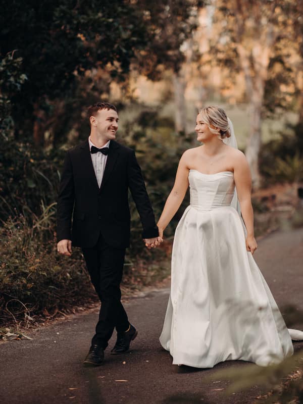 Bride Libby and groom Kyle walk hand in hand along a paved path surrounded by greenery at The Tides during their couple portraits session.