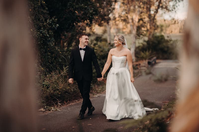Bride Libby and groom Kyle walk hand in hand along a paved path surrounded by greenery at The Tides during their couple portraits session.