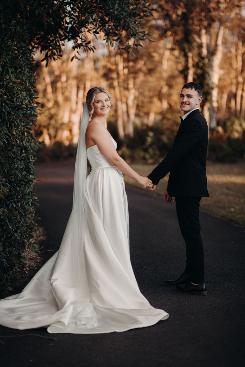 Bride Libby and groom Kyle hold hands and face the camera while standing on a paved path surrounded by greenery and trees. Engagement ceremony by She Said Yes.