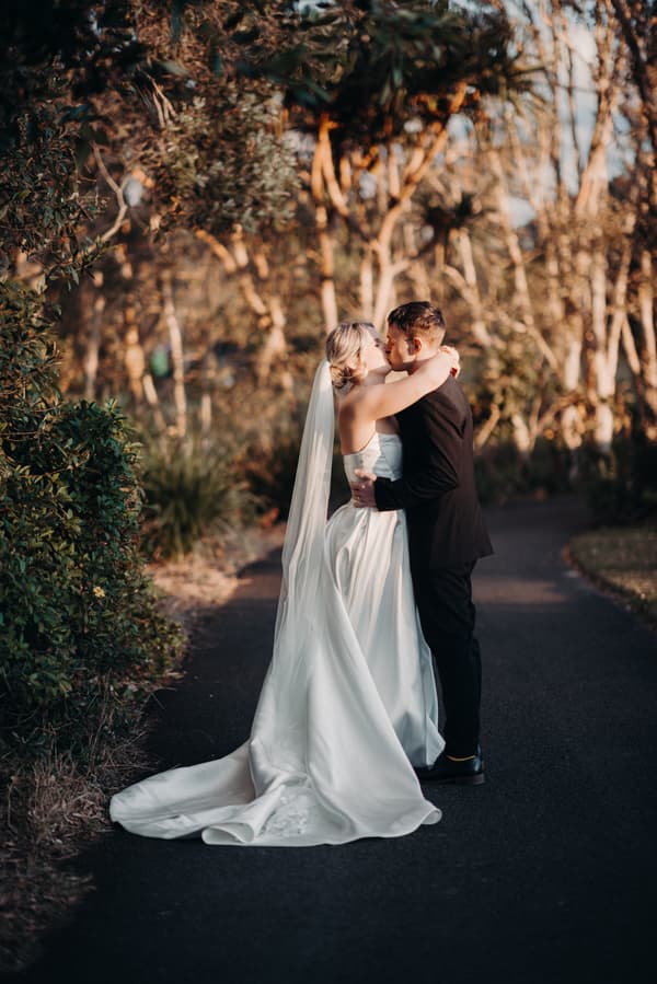 Bride Libby and groom Kyle embrace and kiss on a paved path surrounded by trees at The Tides during their couple portraits session.