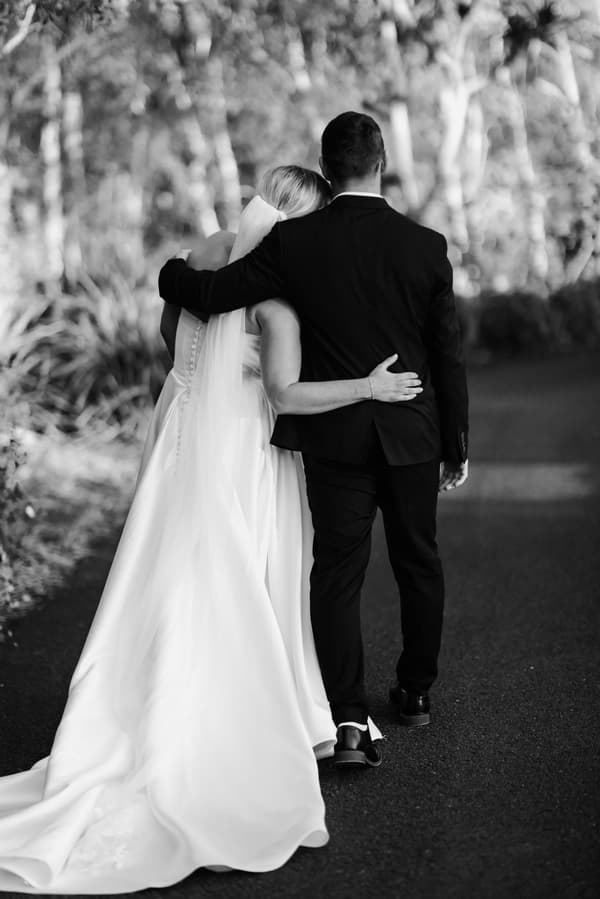 The bride Libby and groom Kyle walk arm in arm down a path at The Tides, seen from behind. Libby wears a long white wedding gown with a veil, and Kyle wears a dark suit.