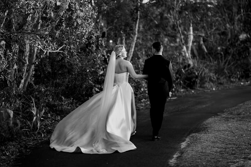 Bride Libby and groom Kyle walk together on a paved path surrounded by dense trees at The Tides, with Libby holding onto Kyle's arm and her long veil and dress trailing behind her.