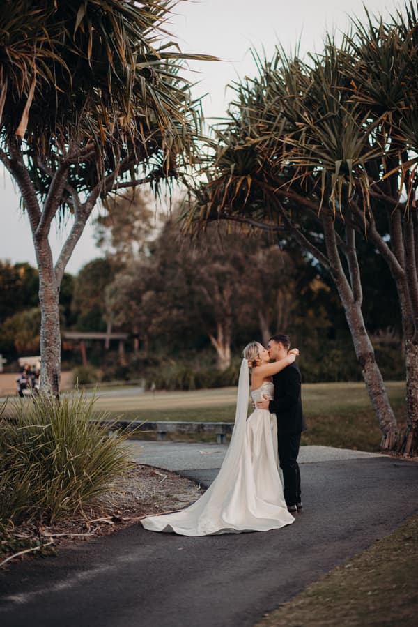 Bride Libby and groom Kyle embrace and kiss on a paved path surrounded by trees and greenery at The Tides.