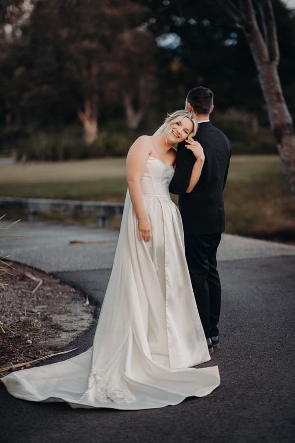Bride Libby in a white wedding gown leans on groom Kyle's shoulder as he stands with his back to the camera on a paved path at The Tides.