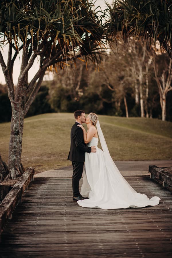 Bride Libby and groom Kyle share a kiss on a wooden bridge at The Tides, surrounded by trees and grassy hills.