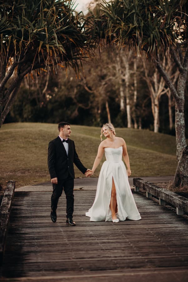 Libby and Kyle walk hand in hand on a wooden bridge at The Tides, surrounded by trees and grassy hills.
