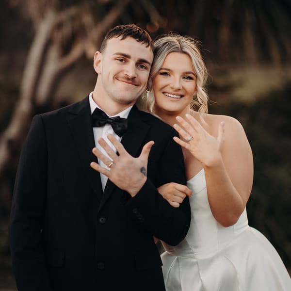 Bride Libby and groom Kyle pose together at The Tides, showing their wedding rings to the camera. Libby wears a white strapless wedding gown and Kyle wears a black tuxedo with a bow tie.