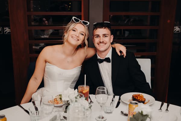 The bride Libby and groom Kyle sit together at a reception table in The Tides — The Pandanus Room, smiling at the camera with plates of food and drinks in front of them.