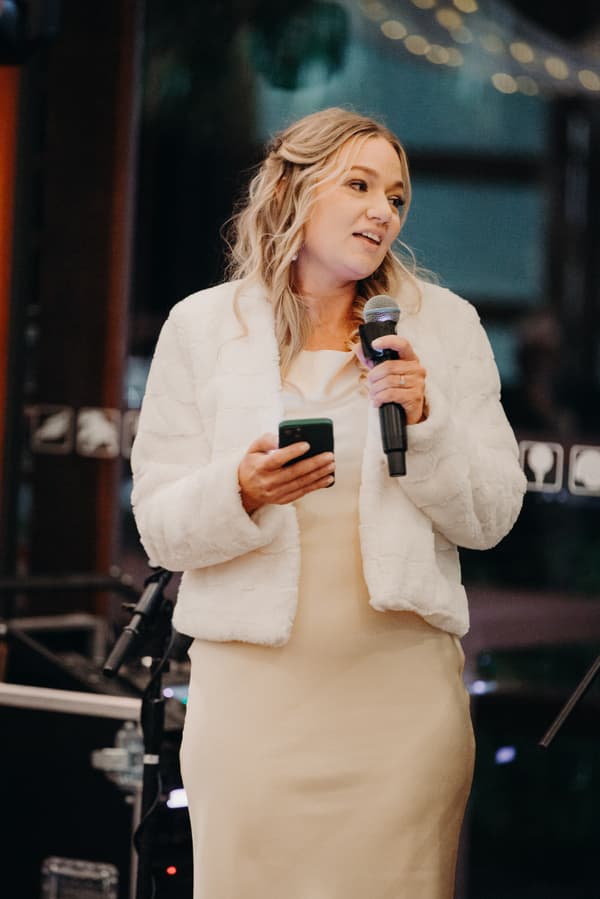 The bride Libby stands in The Pandanus Room at The Tides reception, holding a microphone and a phone while speaking.