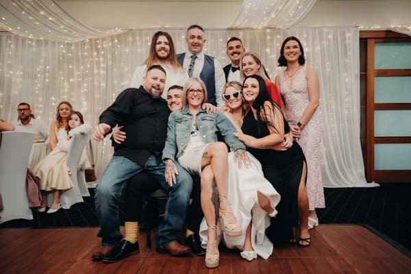 A group of wedding guests pose together in The Pandanus Room at The Tides during the reception. The group includes men and women, some seated and some standing, with a backdrop of white drapes and string lights. Additional guests are seated at tables in the background.