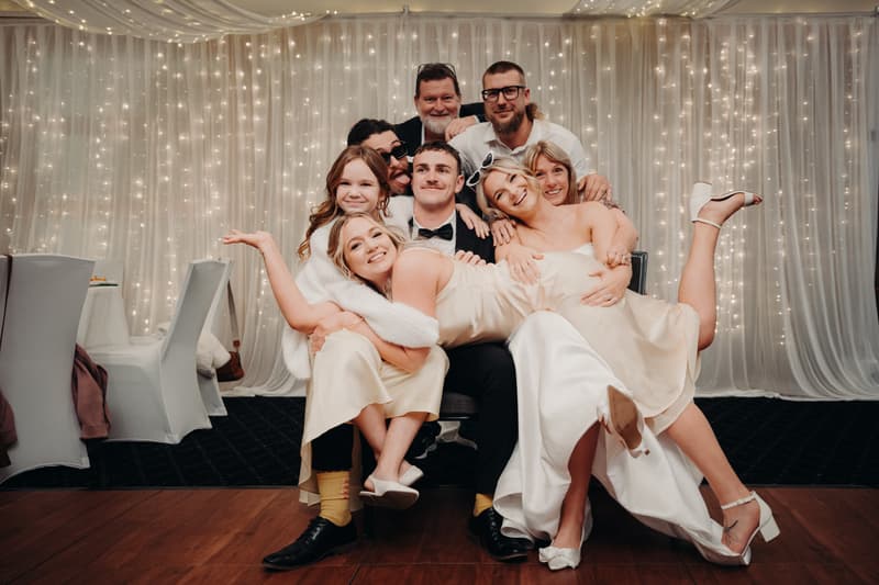 Kyle and Libby pose with six guests in The Pandanus Room at The Tides during the wedding reception, with Libby in a white gown and Kyle in a black suit seated in the center, surrounded by guests in light-colored attire against a backdrop of white curtains and string lights.