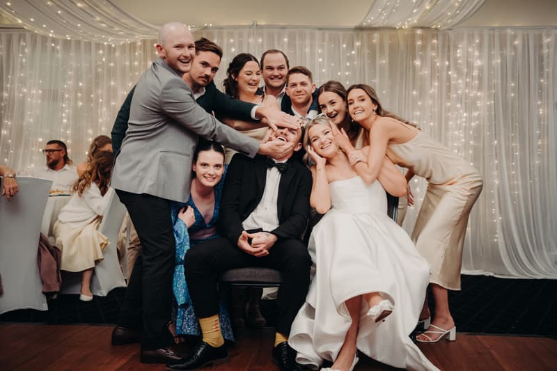 The bride Libby in a white gown and the groom Kyle in a black tuxedo sit on chairs surrounded by eight wedding guests posing playfully in The Pandanus Room at The Tides reception.